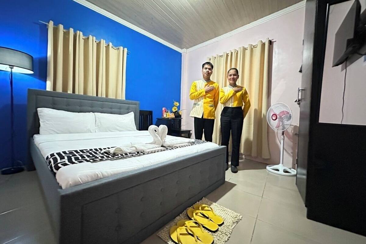 Une chambre avec un lit, deux personnes portant des t-shirts jaunes, un ventilateur et un tapis avec des sandales.