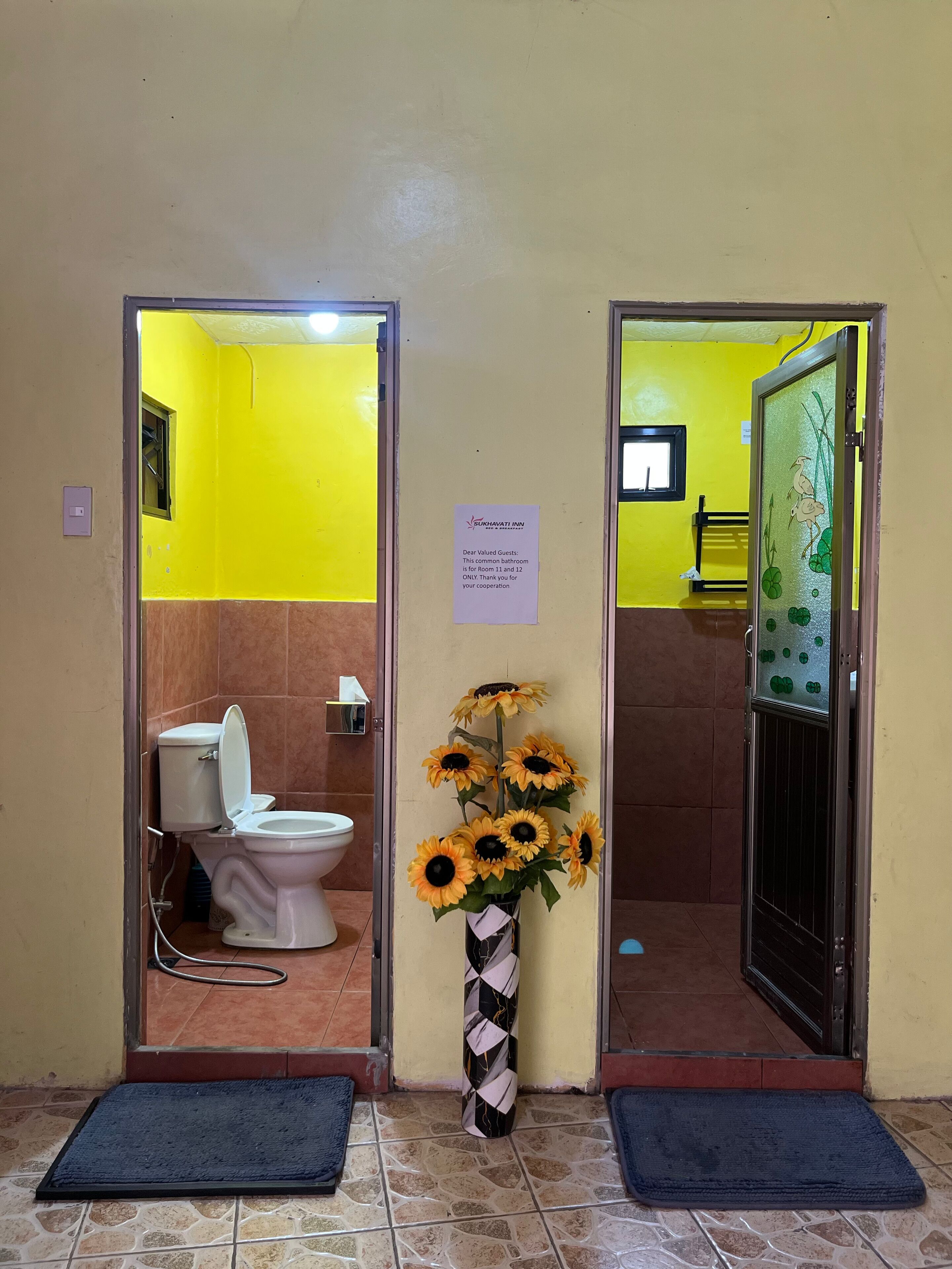 A bathroom with a toilet, a sink, and a mirror. There is a sign on the wall and a vase with sunflowers.