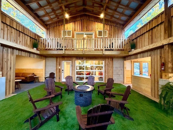 A wooden cabin with a central fire pit, multiple Adirondack chairs, and a balcony overlooking a grassy area.