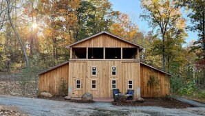 A wooden barn-style building with a large upper level, a gravel driveway, and surrounding trees.