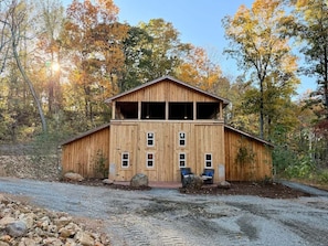 A wooden barn-style building with a large upper level, a gravel driveway, and surrounding trees.
