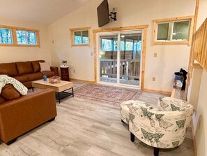 A living room with a brown sofa, a patterned armchair, a coffee table, and a large sliding glass door leading to a deck.