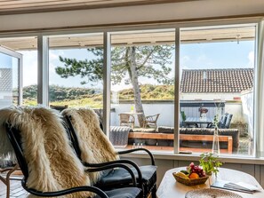 Ett mysigt rum med ett stort fönster, ett träpanelat tak, en fåtölj klädd i fäll, ett bord med en fruktskål och utsikt över ett hus och ett landskap.