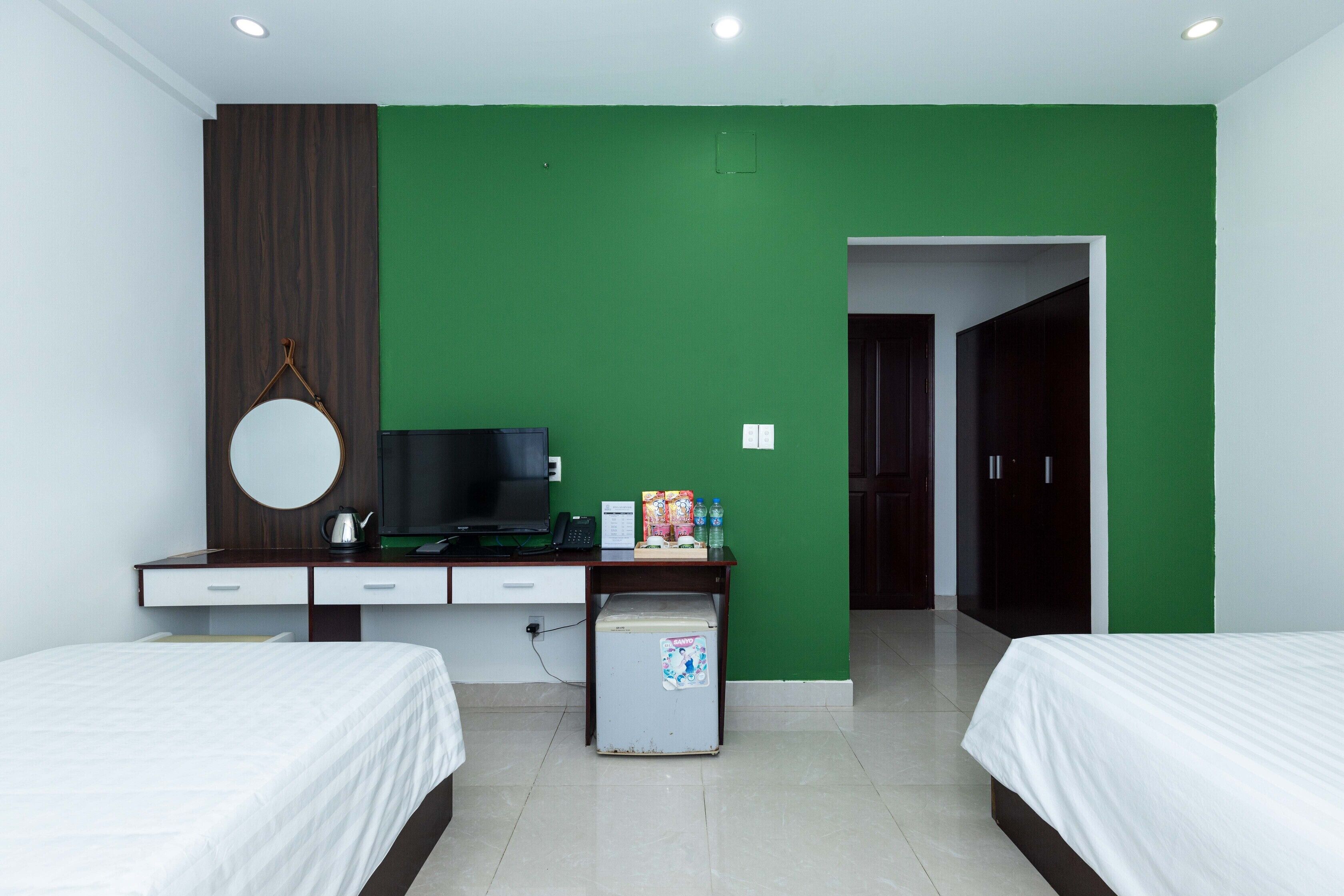 A hotel room with two beds, a desk with a television, a small refrigerator, and a green accent wall.