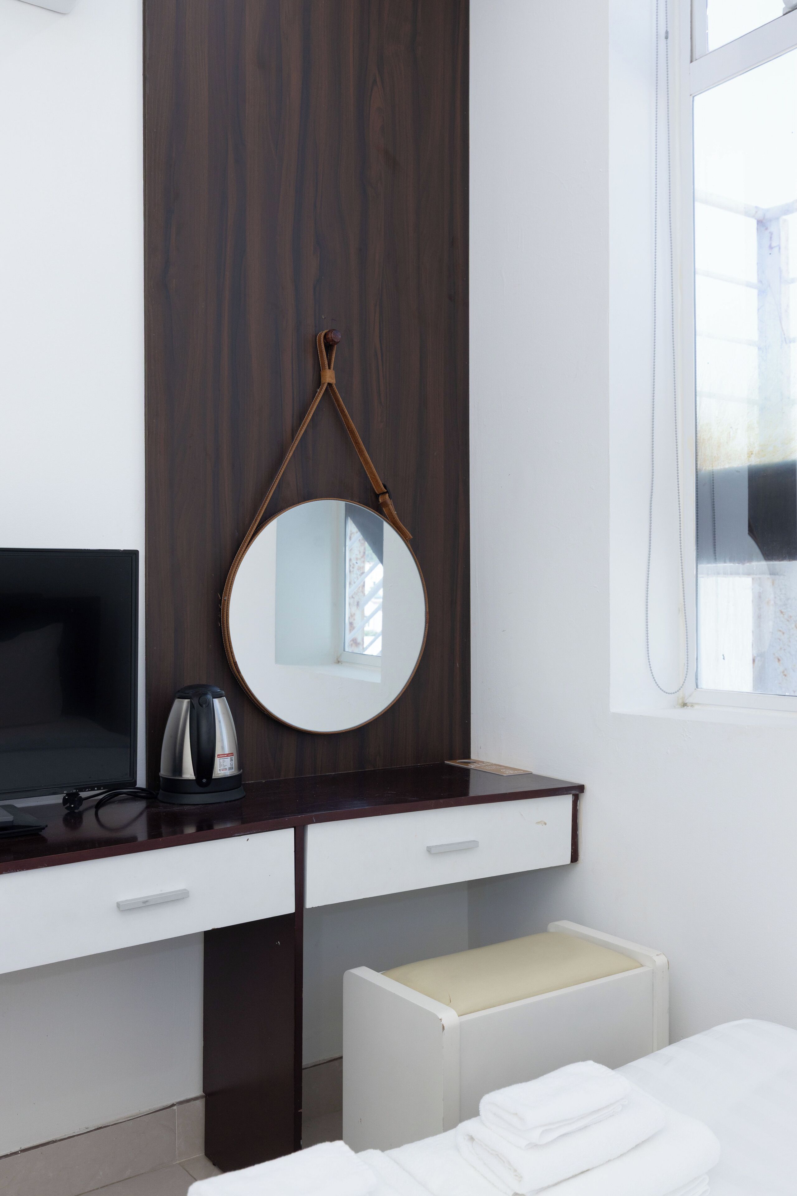 A modern hotel room with a dark wood panel wall, a round mirror, a flat-screen TV, a coffee maker, a white bedside table, and a window with a view.