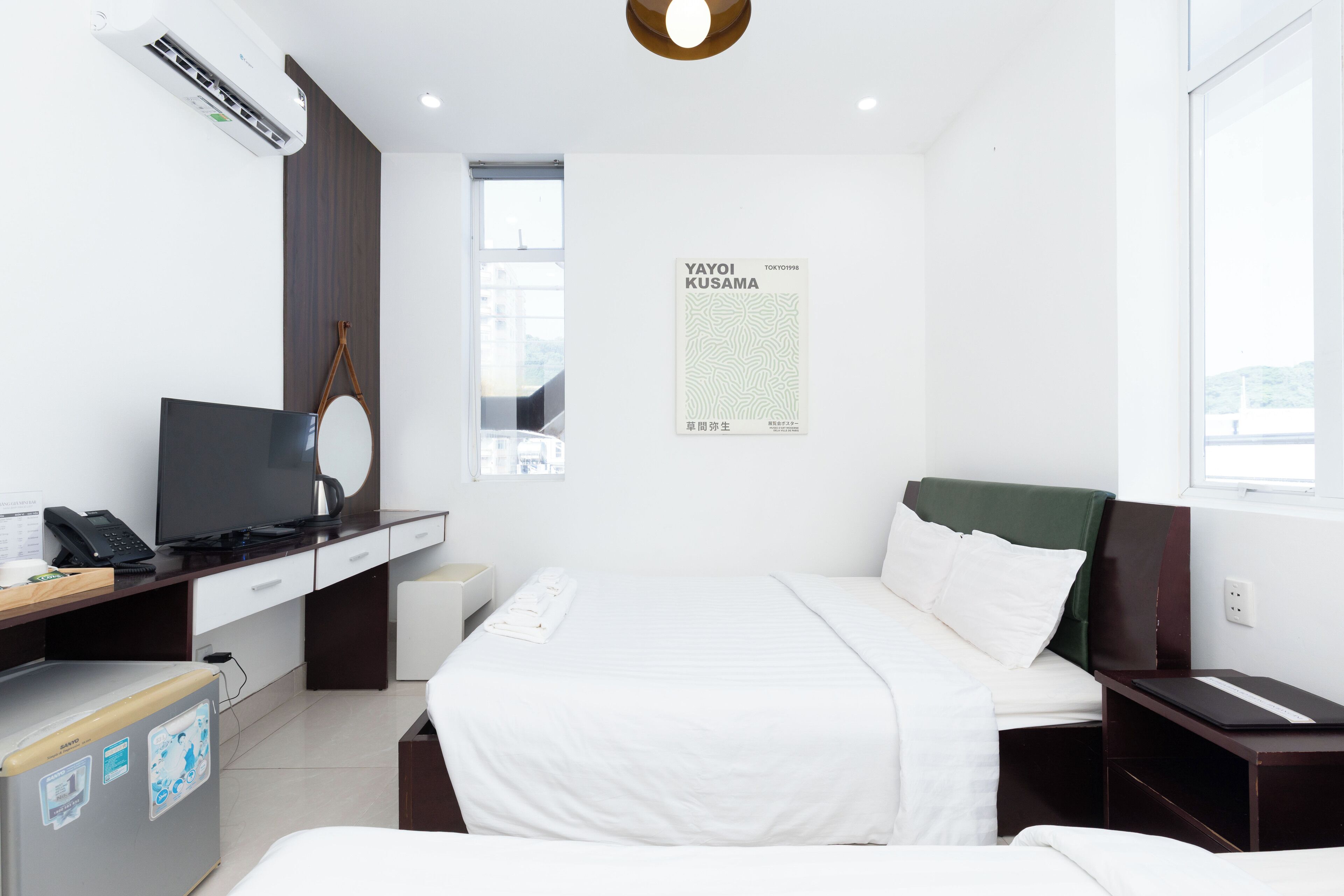 A hotel room with a bed, a desk with a computer and phone, a mirror, and a poster on the wall.