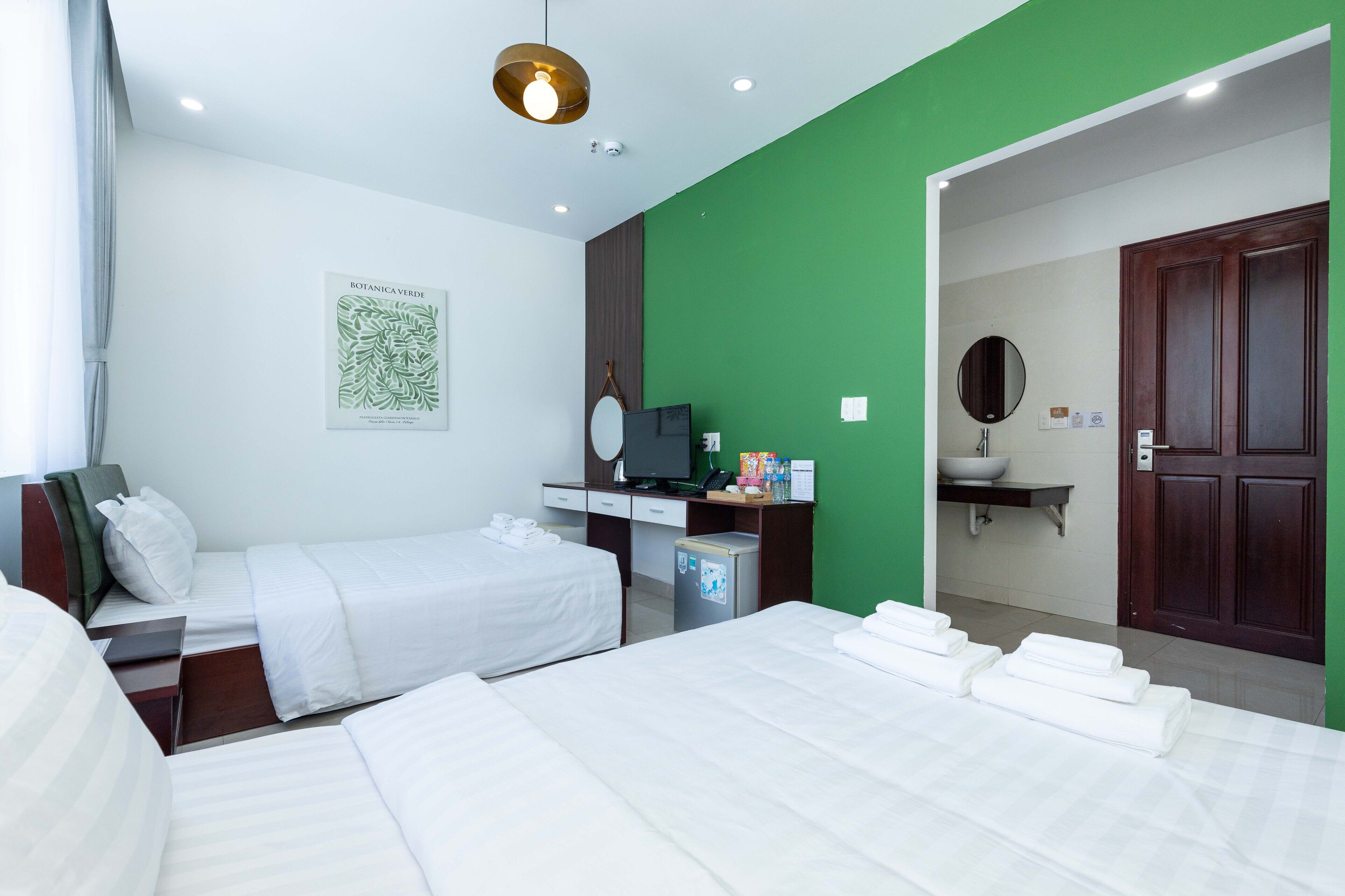 A hotel room with two beds, a desk with a computer, a green accent wall, a mirror, and a door leading to a bathroom.