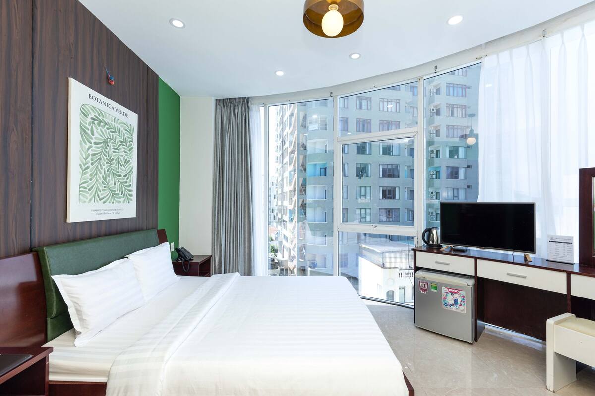 Ein Hotelzimmer mit einem großen Bett, einem Schreibtisch mit Fernseher und Blick auf eine Stadtlandschaft durch große Fenster.