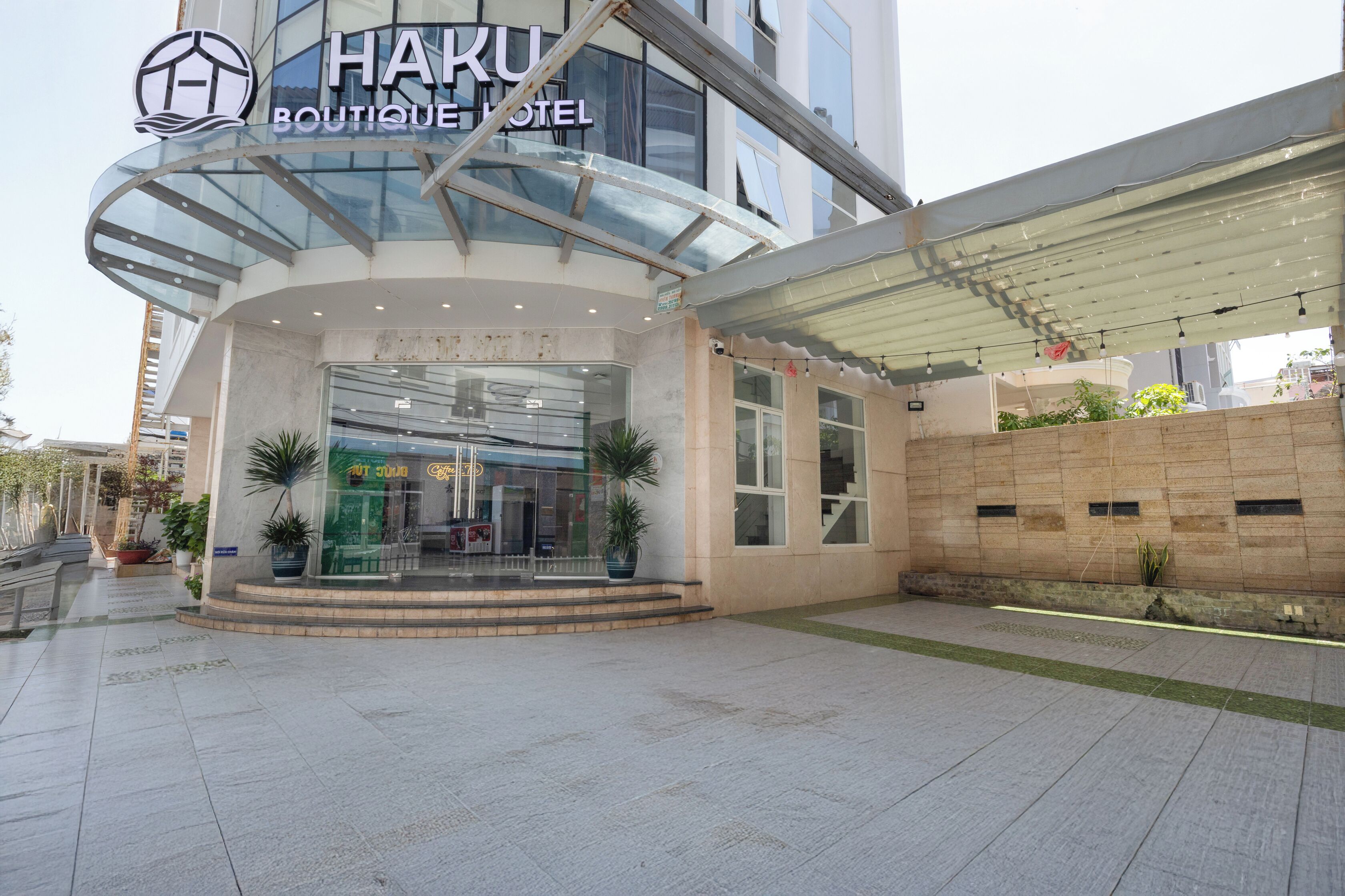Entrada moderna de hotel com fachada de vidro, área de acesso coberta e plantas em vasos.