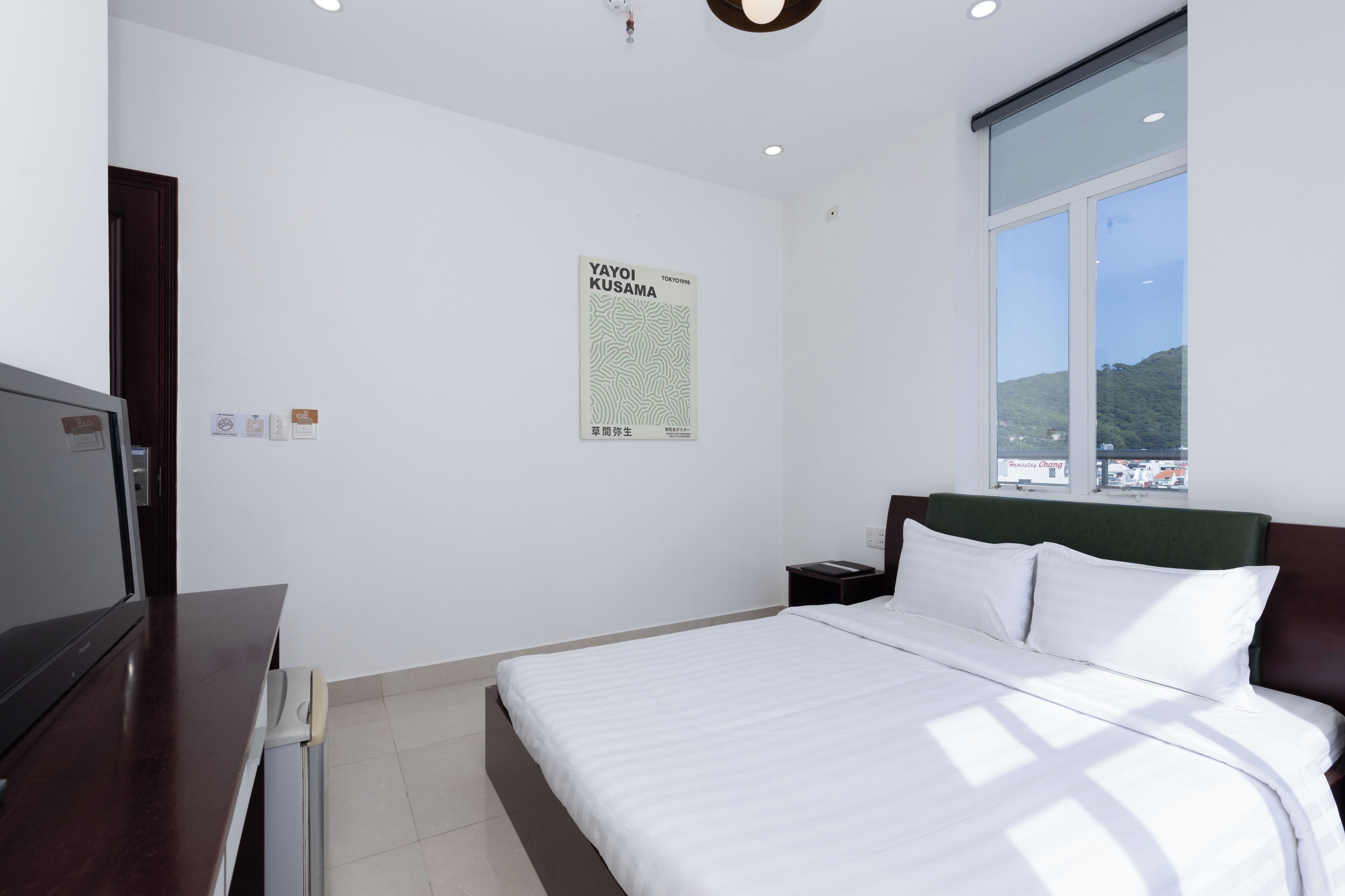 A hotel room with a bed, a desk, a TV, a poster on the wall, and a window with a view of hills.