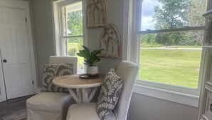 A room with two white cushioned chairs, a small round table with a potted plant, and a view of a grassy yard through large windows.
