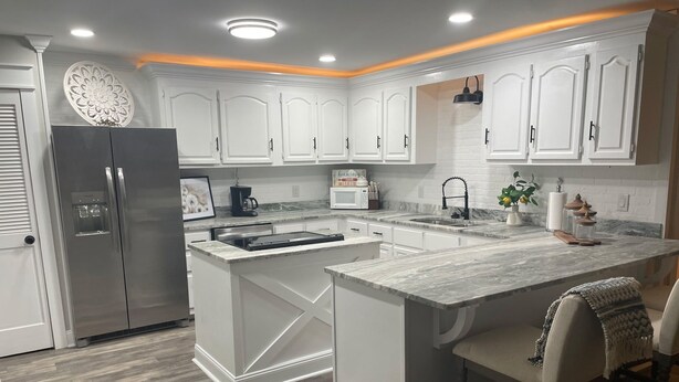 A modern kitchen with white cabinetry, a stainless steel refrigerator, a central island with a sink, and a bar area with stools.