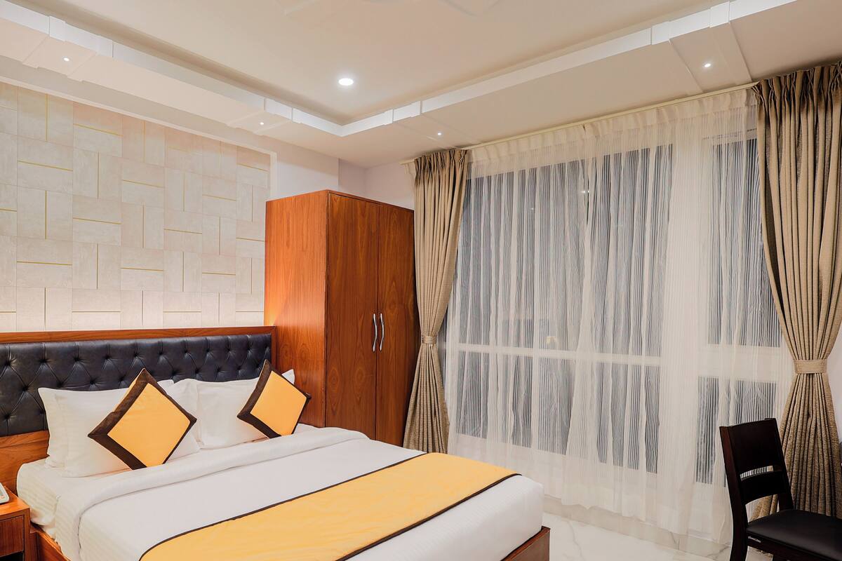Una habitación de hotel con una cama grande, un armario de madera, una mesita de noche con lámpara y teléfono, y una ventana con cortinas translúcidas.