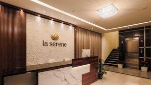 Un comptoir d’accueil avec un plateau en marbre, un panneau décoratif en bois et un logo au-dessus du nom « La Sérène ». Un escalier mène à un ascenseur, et des plantes en pot sont disposées près du comptoir.
