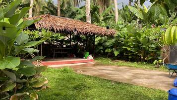 A thatched-roof structure with a concrete platform, surrounded by lush greenery and palm trees.