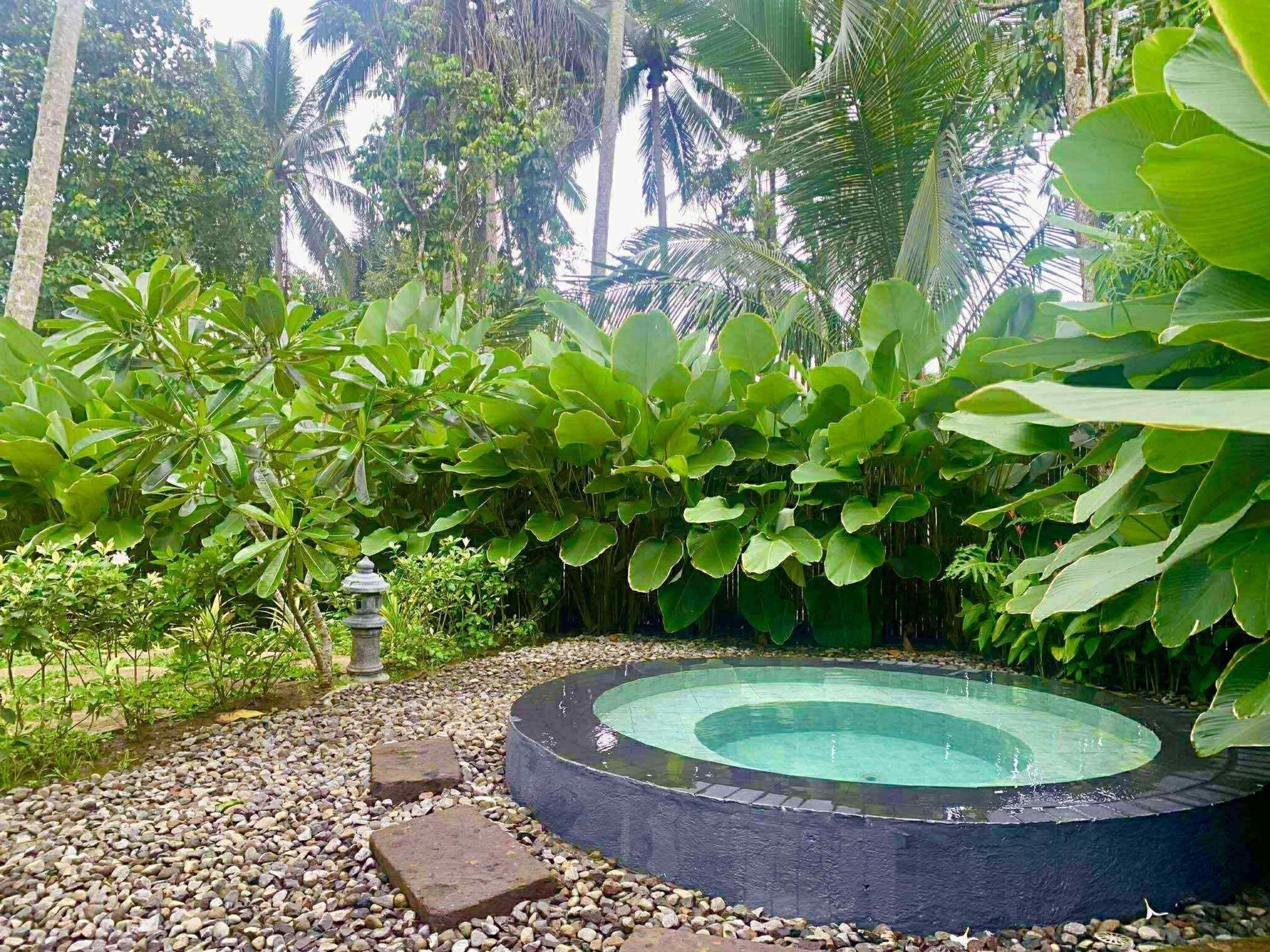 Una piscina circular rodeada de guijarros y exuberante vegetación, con un farol de piedra cerca.