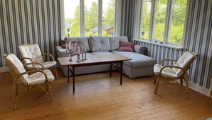 A room with a sectional sofa, two wicker chairs, a wooden table, and large windows with a view of greenery outside. - 4 Star Holiday Home in Torsaker (Torsåker)