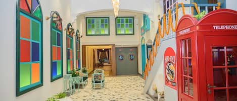 Un vestíbulo de hotel con una escalera central, un teléfono rojo y diseños coloridos en las ventanas.