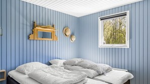 Une pièce aux murs bleus, un lit avec une literie blanche, une fenêtre donnant sur des arbres et une étagère en bois où sont posés des chapeaux.