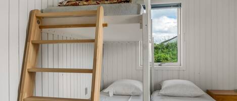 A bunk bed room with a wooden ladder, two beds, a window with a view of greenery, and a small wooden table.