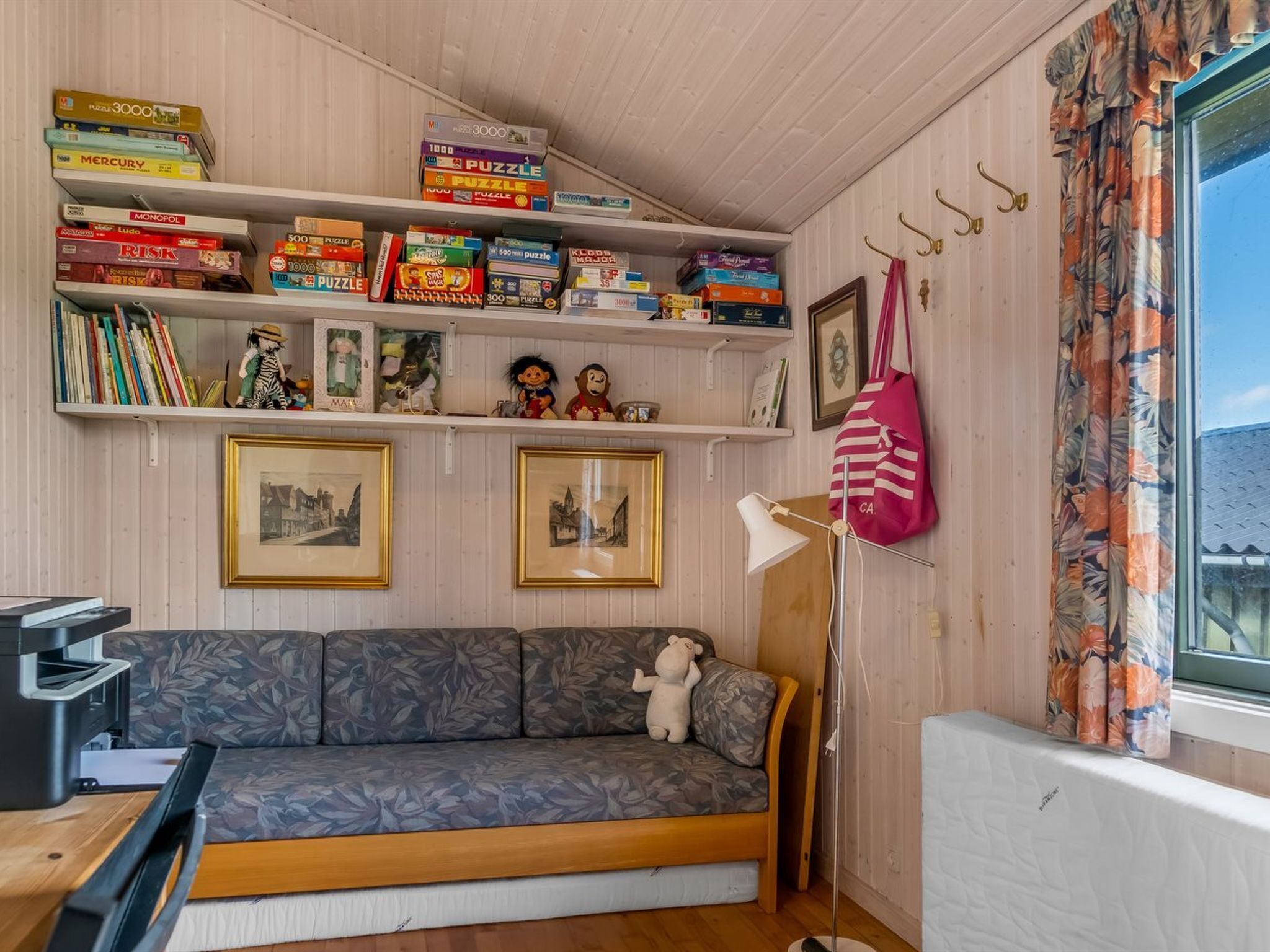 A cozy room with a sofa, a bookshelf filled with board games, framed pictures, and a window with curtains.