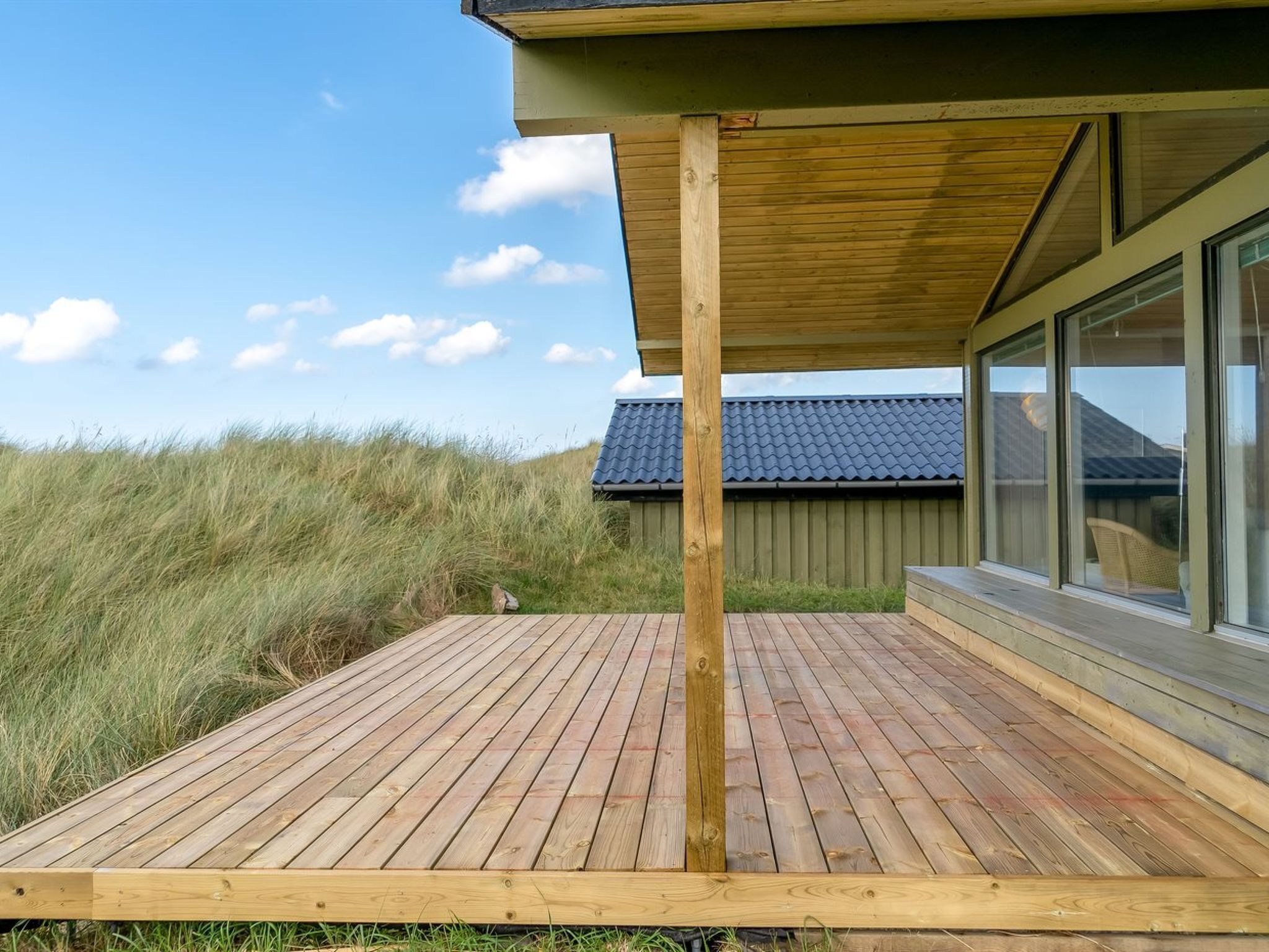 Une terrasse en bois avec un poteau en bois, une fenêtre donnant sur une maison et, au fond, un champ verdoyant.