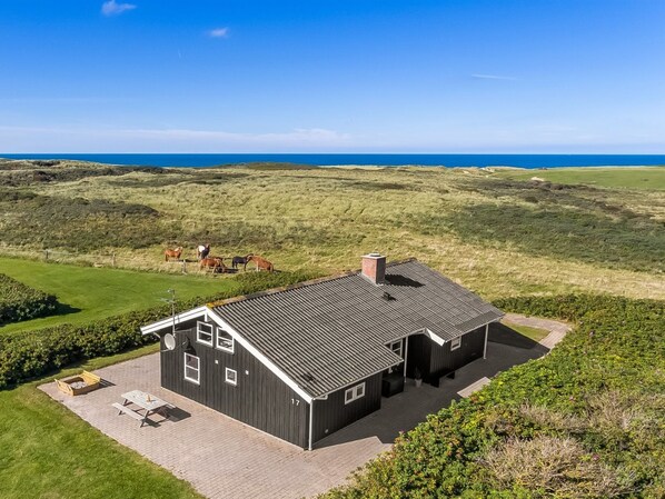 Une maison à deux étages, avec un toit sombre et des éléments décoratifs blancs, entourée d’une pelouse bien entretenue et d’un champ où paissent des chevaux.