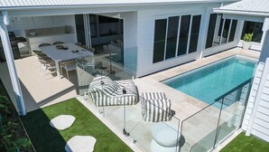 Una casa moderna con un porche de cristal, piscina y zona de comedor con mobiliario exterior.