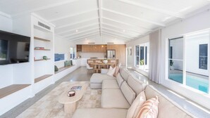 Amplia sala de estar con un sofá grande, una mesa de centro y un comedor con una mesa y sillas de madera. Hay un televisor montado en la pared y un amplio ventanal con vistas al exterior.