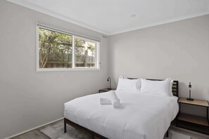 A bedroom with a large window, a bed with white linens, a bedside table, and a nightstand.