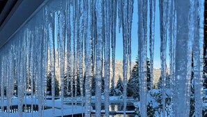 Des glaçons pendent de la rambarde d’un balcon, une terrasse recouverte de neige, et une vue sur un paysage enneigé avec des arbres et un bâtiment en arrière-plan.