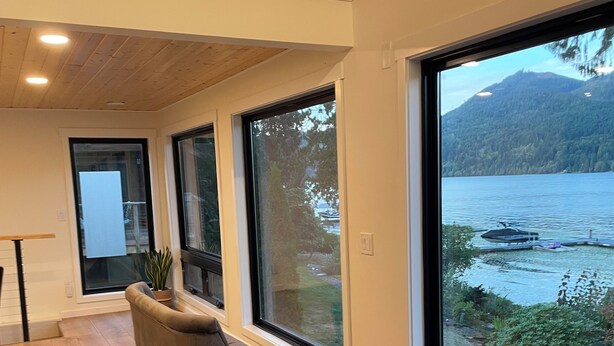 Un salon moderne doté d’une grande fenêtre donnant sur un lac et des montagnes. On y trouve un canapé, un fauteuil et une petite table sur laquelle est posée une couverture.