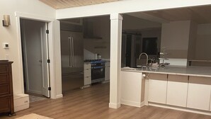 Un vaste espace de vie doté d’un plancher en bois, d’un canapé beige et d’un coin cuisine avec des meubles blancs et un réfrigérateur en acier inoxydable.