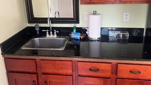 A kitchen with a black countertop, wooden cabinets, a stainless steel sink, a mirror, and a paper towel dispenser. - Kona Nook - Cozy Studio with Island Warmth (Kailua-Kona)