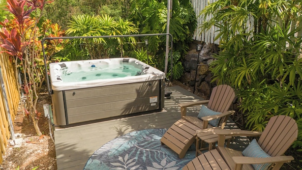 A hot tub surrounded by wooden deck chairs on a patterned rug, with lush tropical plants and a stone wall in the background. - Kona Nook - Cozy Studio with Island Warmth (Kailua-Kona)