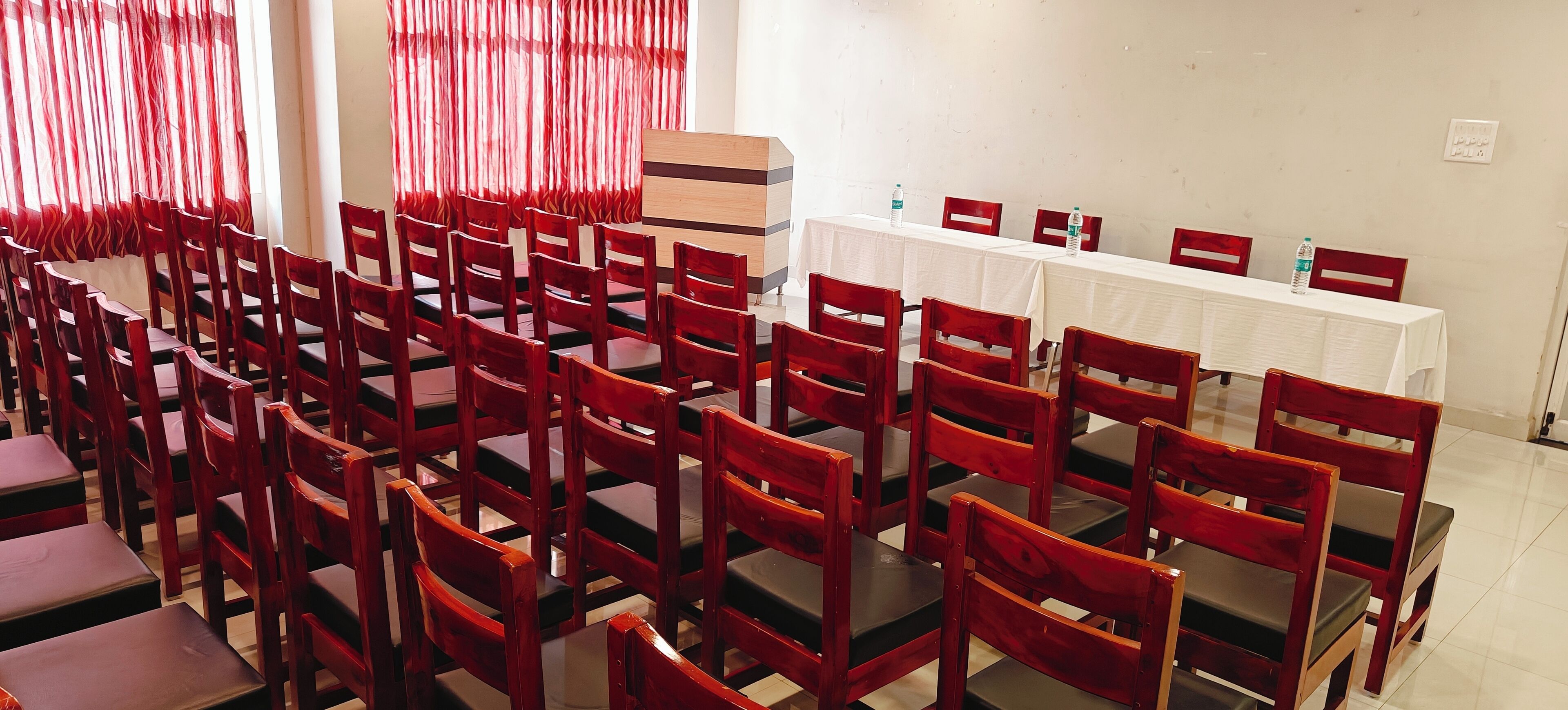 A conference room with rows of wooden chairs and tables, red curtains, and a whiteboard.