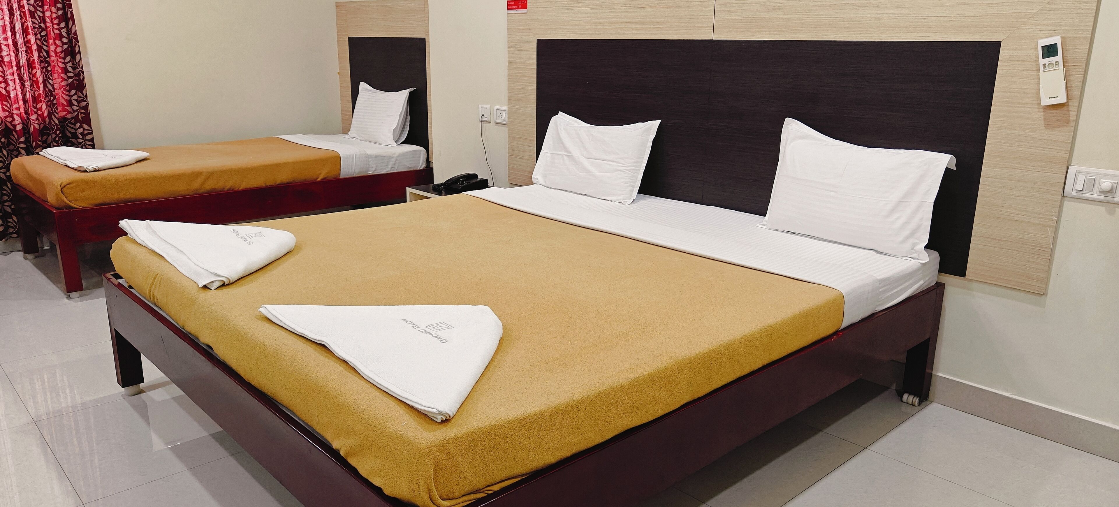 Habitación de hotel con dos camas, cada una con colchón marrón y ropa de cama blanca. Hay una mesita de noche con un teléfono y un termostato en la pared.
