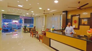 Uma área de recepção de hotel com recepcionista, balcão e um espaço de espera com cadeiras e uma mesa pequena.