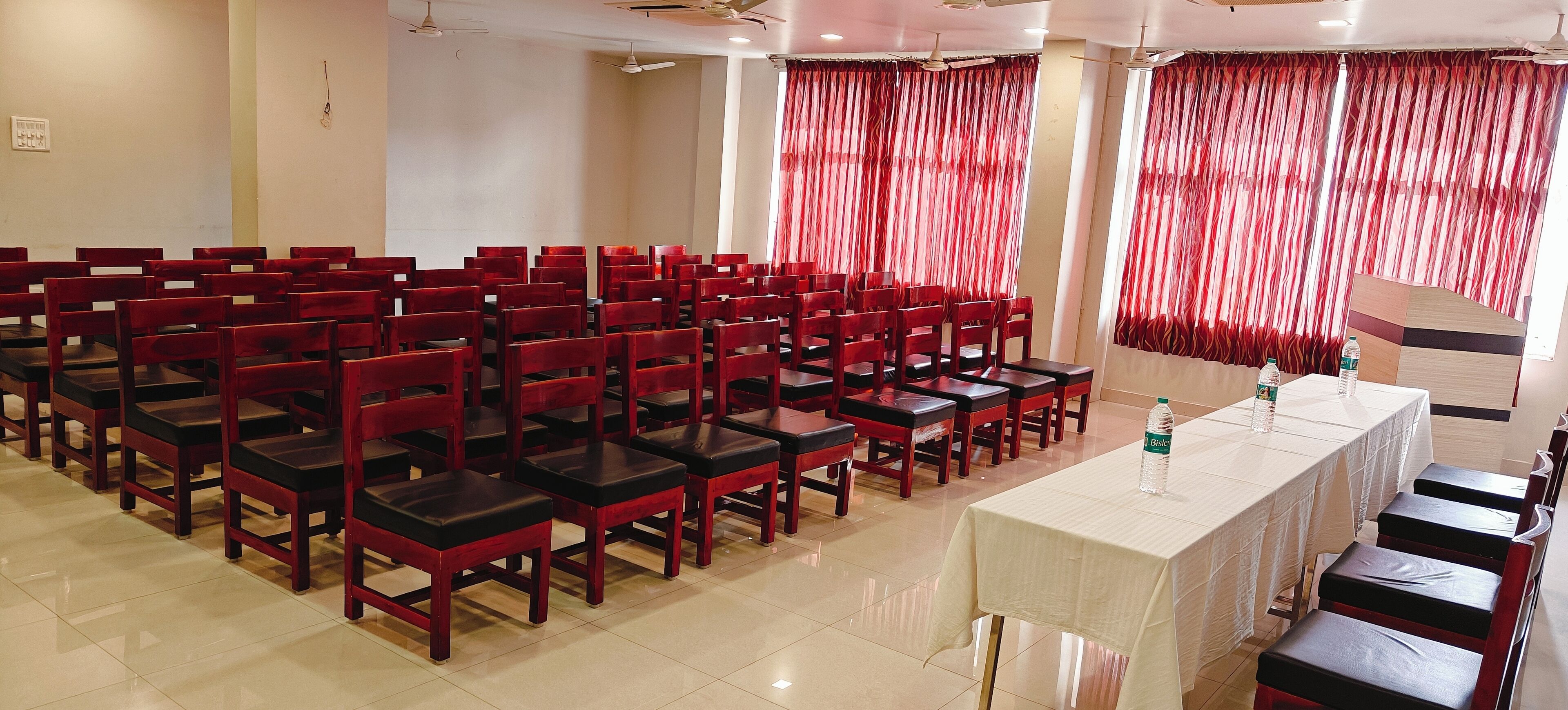 Un salón de conferencias con filas de sillas y mesas de madera, cortinas rojas y ventiladores de techo.