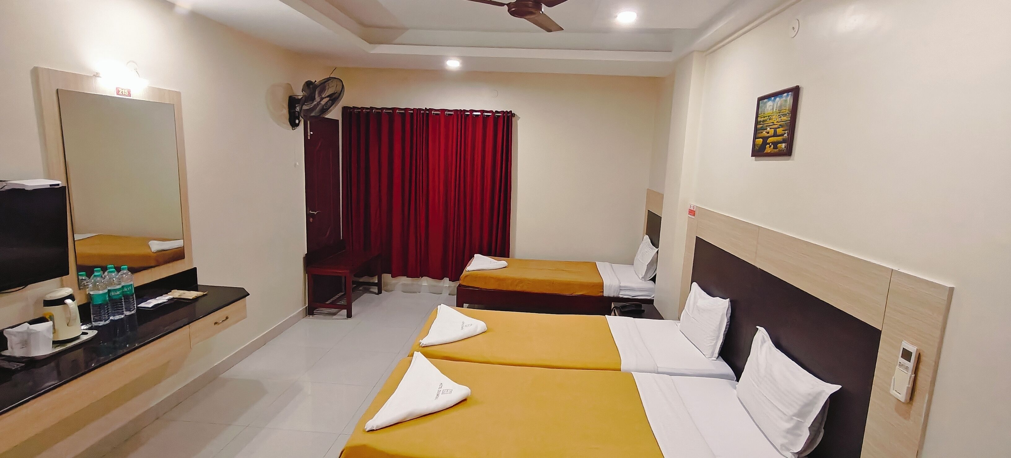 A hotel room with two beds, a TV, a mirror, a red curtain, and a ceiling fan.