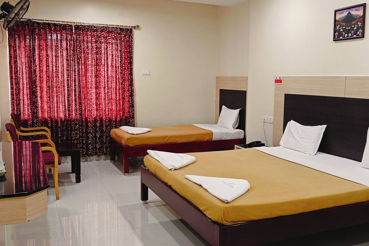 A hotel room with two beds, a desk with a kettle, a chair, and a red curtain.