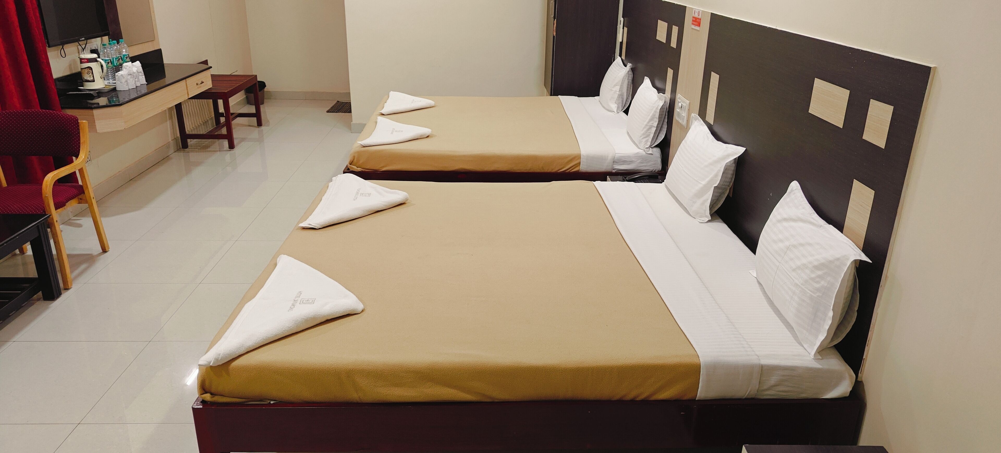 Een hotelkamer met twee bedden, elk met een beige sprei en witte kussens. Er is een bureau met een stoel en een televisie aan de muur.
