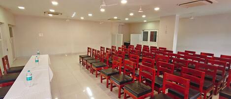 Un salón de conferencias con filas de sillas rojas, una larga mesa con botellas de agua y ventiladores de techo.