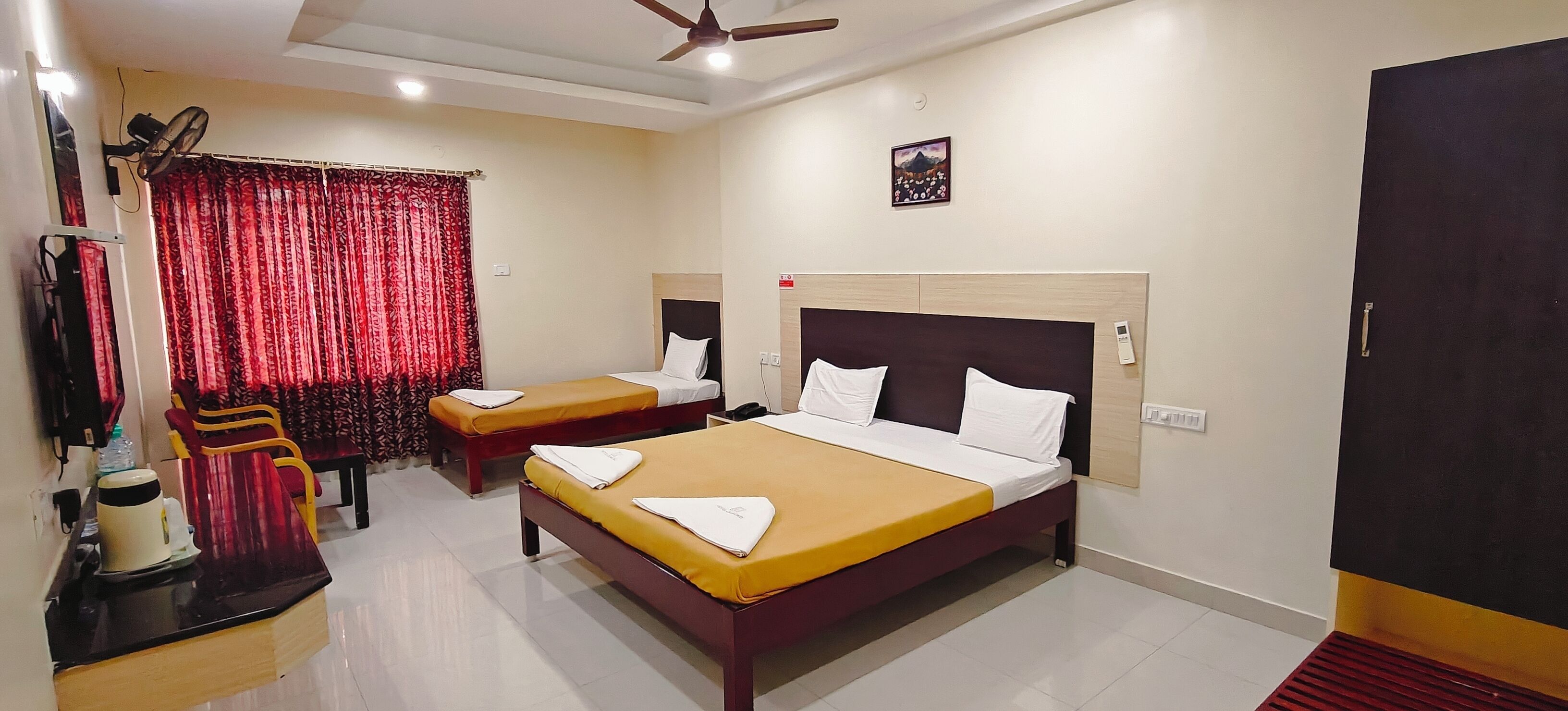 A hotel room with two beds, a desk with a kettle, a fan, and a red curtain.