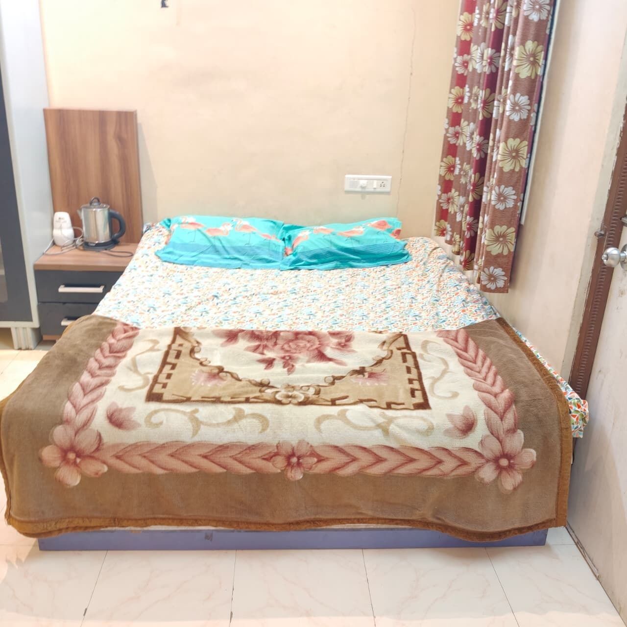 A bed with a floral bedspread and a decorative throw blanket, a wooden nightstand with a kettle, and a floral curtain.