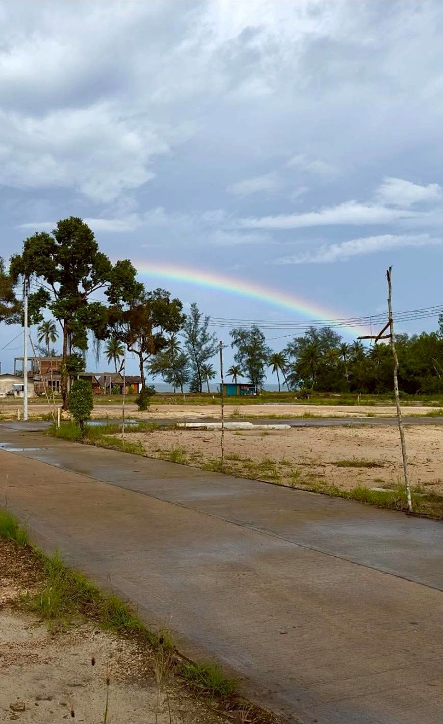 Una carretera pavimentada con césped y árboles, cielo despejado y un arcoíris a lo lejos.