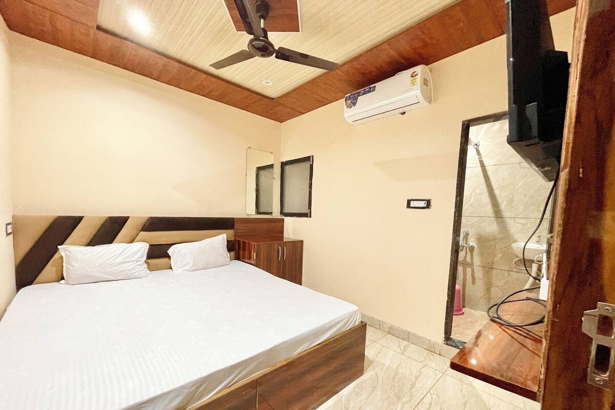 A bedroom with a bed, wooden headboard, ceiling fan, air conditioning unit, and a bathroom with a shower and sink.