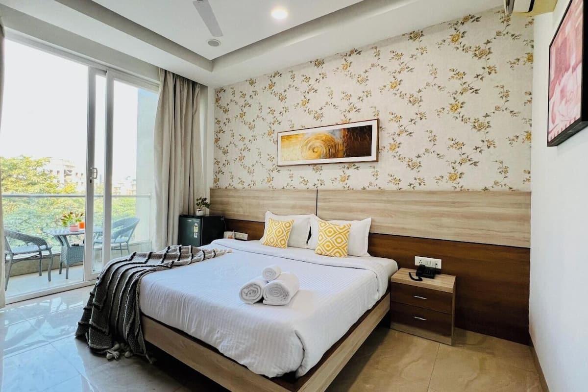 A hotel room with a large bed, a bedside table, a nightstand, a window with curtains, a balcony with outdoor furniture, and a floral wallpaper pattern.