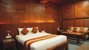 A hotel room with a wooden wall panel design, a bed with white bedding and brown accents, a wooden nightstand, a bedside table with a lamp, and a cushioned armchair. - ila Comfort Harkapur (Devachuli)
