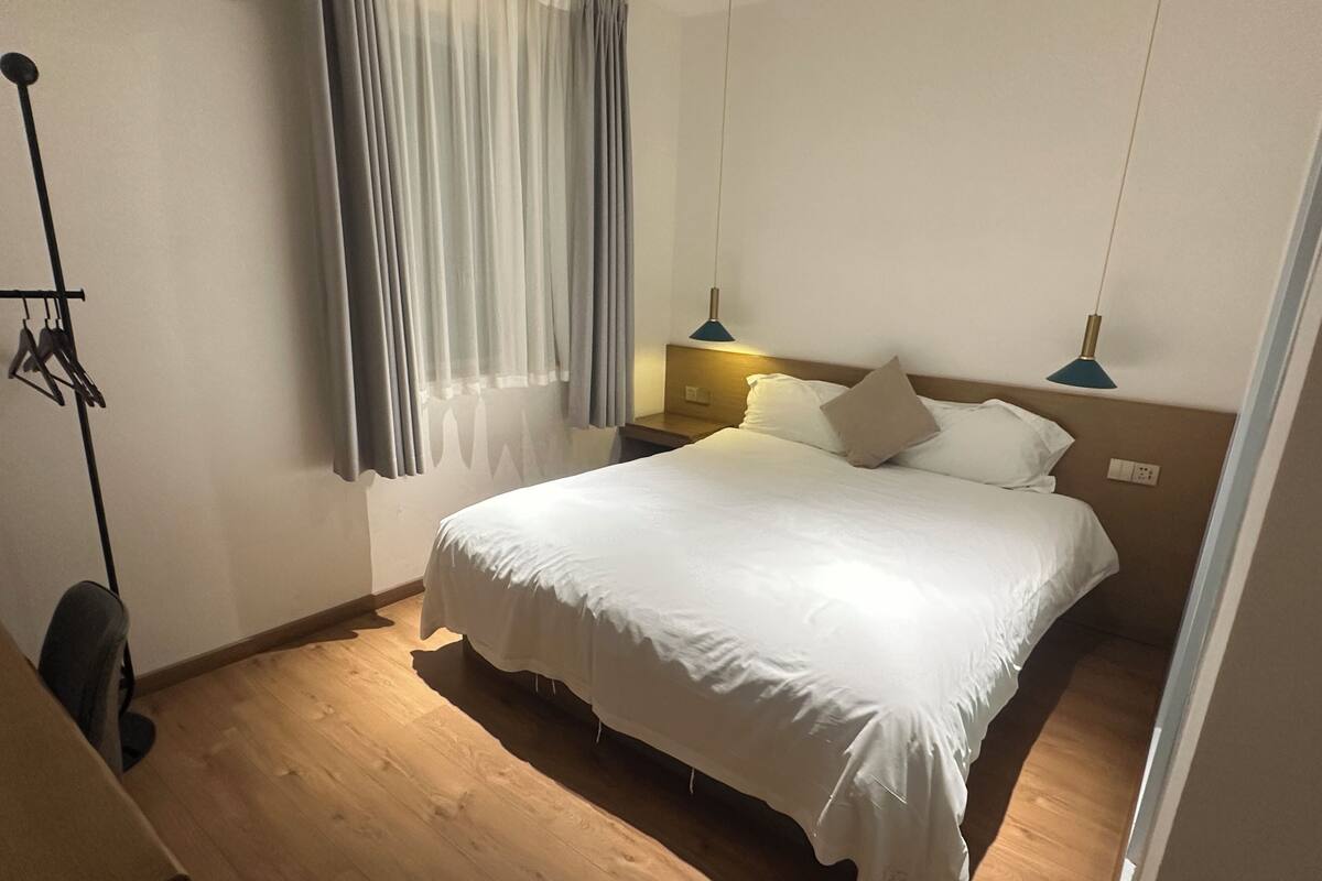 A hotel room with a large bed, a desk, and a chair. There are two bedside lamps and a window with curtains.